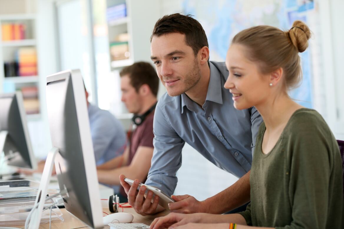 A manager training an employee, both looking at a desktop computer monitor. 
