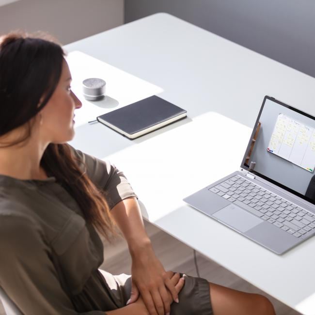 A woman sits at a desk and watches a video on her laptop of a lecturer pointing at a whiteboard.