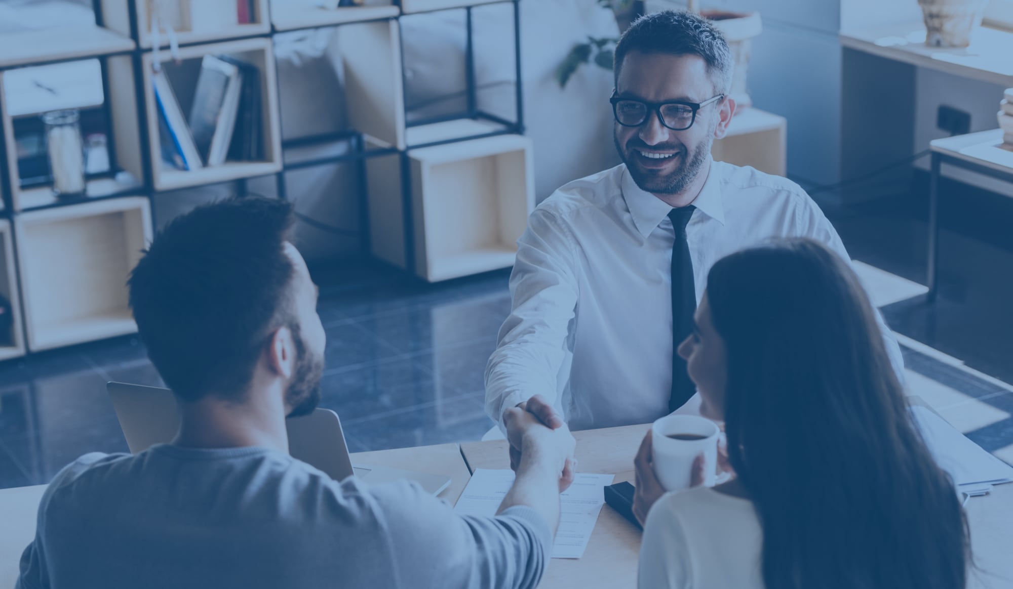 A banker sits across from two clients, shaking hands with one of them.